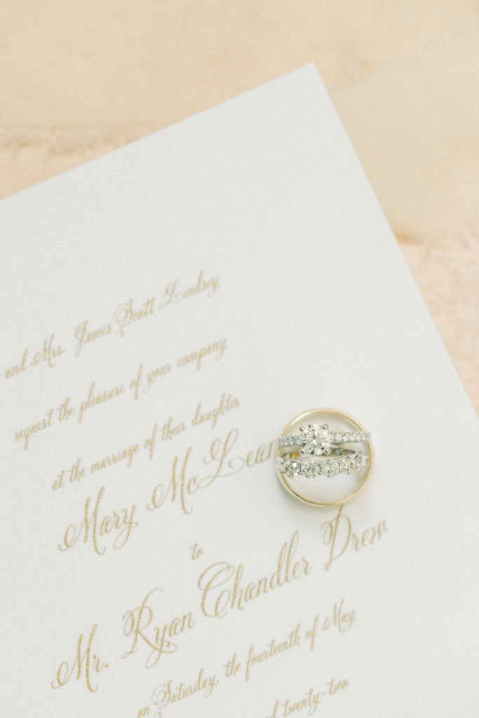 wedding band and engagement ring sit on top of white and gold wedding invitation
