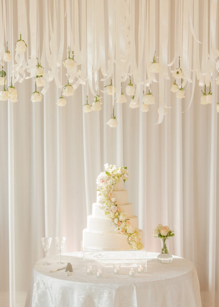 five tier wedding cake sits on a white table with dozens of floating roses above